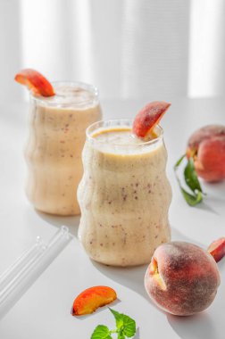 İki bardak şeftalili ve muzlu smoothie pencerenin yanında açık bir arka planda, tropikal meyveler ve sabah güneşiyle birlikte. Sağlıklı beslenme ve detoks diyeti kavramı. Ön görünüm, boşluğu kopyala.