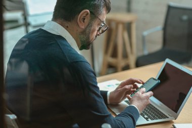 Busy man texting by smartphone while using laptop, sitting in the office