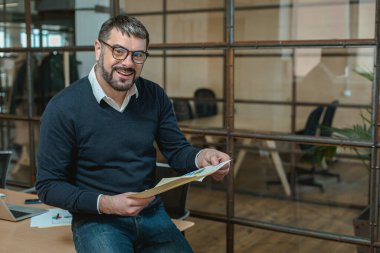 Mutlu yetişkin adam işyerinde dururken belgeleri kontrol ediyor.
