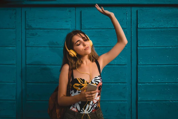 Positive young woman with clothed eyes dancing with raised arm while listening to music in yellow headphones near blue wall