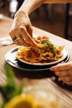 Meçhul bir kadın restoranda masada oturuyor ve üzerine peynir ve guacamole sosu dökülmüş lezzetli nachos yiyor.