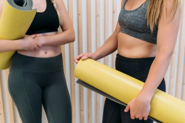 İki atletik genç bayan, sarı yoga minderlerini tutarak fitness derslerine hazırlanıyorlar.