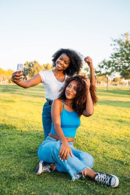 İki arkadaş çimlerin üzerinde oturmuş güneşli bir günde akıllı telefonlarıyla selfie çekiyorlar.