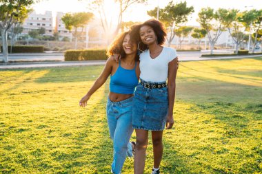 İki arkadaş birlikte parkta yürüyorlar, güneşin altın ışığında gülüyorlar.