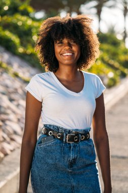 Güneşli bir günde afro saçlı, gülümseyen ve dışarıda yürüyen genç siyahi bir kadın.