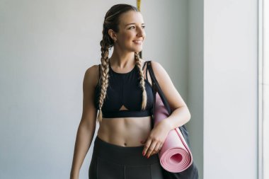 Sportif kadın yoga minderini tutuyor gülümsüyor ve spor salonuna bakıyor.