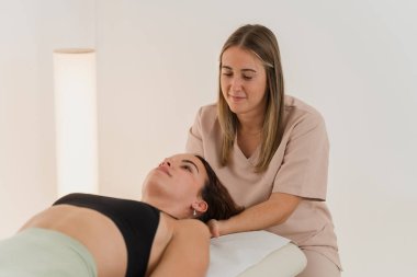 Modern klinikte masaj masasında yatan hastanın boynunu dikkatlice inceleyen kadın fizyoterapist.