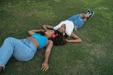 İki genç kadın çimenlerde uzanıyor, kulaklıklarla müzik dinliyor, rahatlama ve dostluk anlarını paylaşıyorlar.