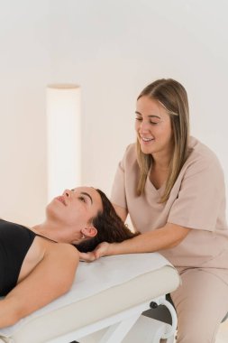 Gülümseyen fizyoterapist masaj masasında uzanan kadının boynunu nazikçe manipüle ediyor.