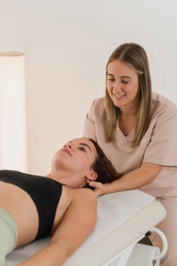 Kadın fizyoterapist masaj masasında uzanan bir kadının boynunu nazikçe manipüle ederek ağrı kesici veriyor ve hareket kabiliyetini geri kazandırıyor.