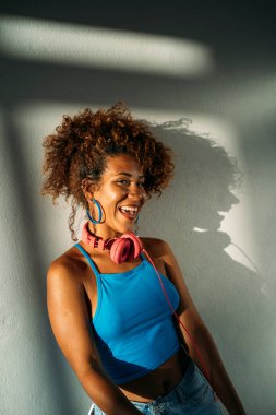 Genç kadın kulaklıkla müzik dinlerken gülümsüyor, pencereden gelen güneş ışığının tadını çıkarıyor.