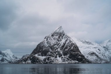 Lofoten adalarındaki Norveç denizinin sakin sularından görkemli bir şekilde yükselen karlı dağları barındıran dramatik bir manzara.