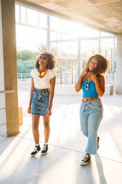 Two young women are happily walking through a sunlit breezeway while wearing headphones clipart