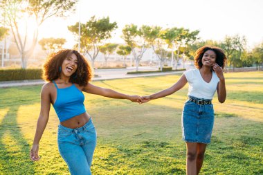 Güneşli bir parkta yürürken iki genç kadın el ele tutuşup gülüyor.