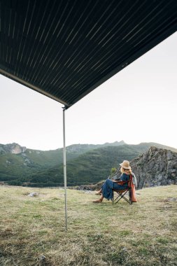 Bir adam, bir sandalyede, bir gölgeliğin altında, huzurlu bir dağ manzarasının tadını çıkarıyor.
