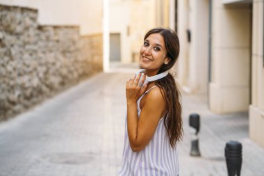 Dar bir sokakta, boynuna kulaklık takıp gülümseyen mutlu genç bir kadın.