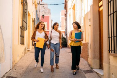 Üç mutlu değişim öğrencisi ellerinde defterlerle dar bir caddede yürüyüp sohbet ediyorlar.