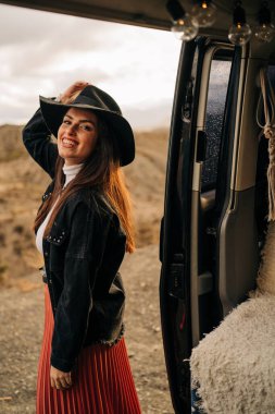 Şapkalı şık bir kadın karavandan iniyor, güzel bir çöl manzarasında minibüs hayatının tadını çıkarıyor.