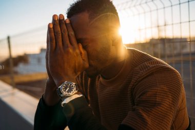 Elleri yüzü kapalı genç bir adam, gün batımında umutla dua ediyor.