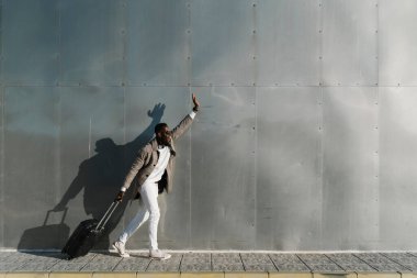 Modern metal bir duvarın yanında bavuluyla yürürken şık bir işadamı el sallıyor.