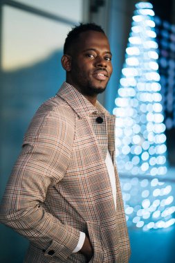 Kendine güvenen siyah girişimci bulanık noel ışıklarının yanında duruyor, şık bir ceket ve beyaz gömlek giyiyor.