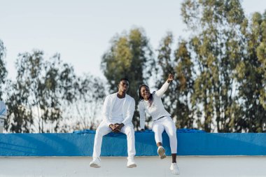 Beyaz elbiseli iki model bir çatıda oturuyor, biri heyecanlı bir şekilde, açık gökyüzüne karşı gösteriş tarzı ve isteklerini sergiliyor.