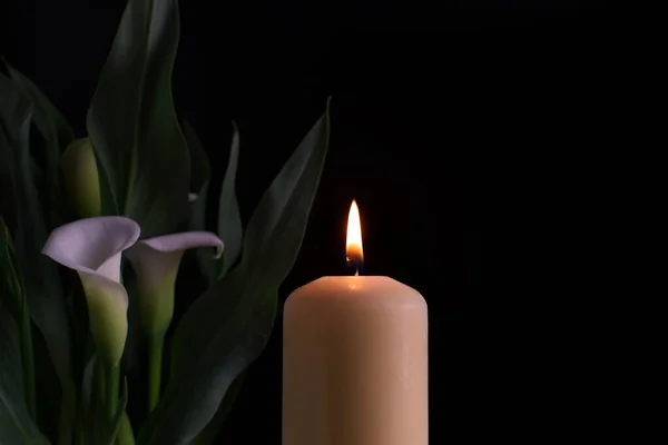 Close up of candle burning flame in the darkness and arum lilies illuminated by the candlelight alongside in a conceptual image