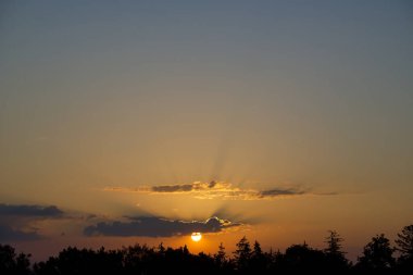 Güneş bir kaç ince bulutla çevrilidir, gökyüzü kahverengiyle kırmızının büyüleyici bir karışımıdır, parlak bir turuncu, sarı ve pembe artçı ışıltıyla aydınlanır.