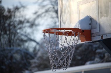 Arka planda bulanık bir basketbol potası. Ağ ve çemberin kenarı bir kar tabakasıyla kaplı. Sahneye eşsiz bir soğuk dokunuş sergiliyor.