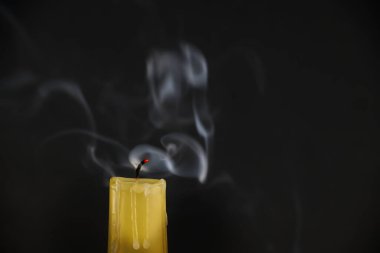 Yellow candle and its wick, which appears red is still emitting smoke after the flame has been extinguished. White smoke is rising in various directions, forming delicate pattern against dark backdrop clipart
