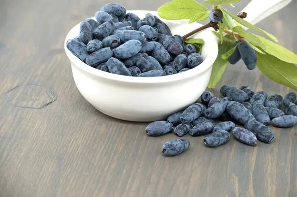 stock image White porcelain bowl filled with blue honeysuckle berries is situated on a wooden table, some of berries spilling out onto the table near branch with fresh green leaves and berries