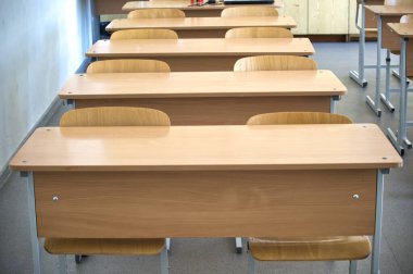 Sıraya dizilmiş tahta masaları ve sandalyeleri olan boş bir sınıfın fotoğrafı, pencerelerden süzülen güneş ışığı.