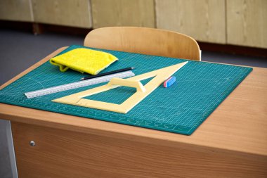 Sınıf masasında cetvel ve kare gibi matematik araçları sergilenirken, bir silgi ve bir kesme paspas üzerindeki temizlik beziyle birlikte.