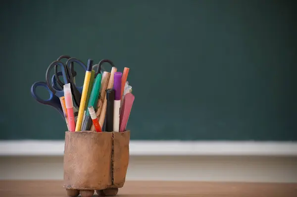 stock image A variety of classroom supplies, including pens, markers, and scissors, organized in a wooden holder in front of a chalkboard.