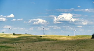 Rüzgâr türbinleri, yeşil ve sarı tarlaları olan yuvarlanan bir arazide duruyor. Mavi gökyüzünün altında, tüylü beyaz bulutlarla süslenmiş..