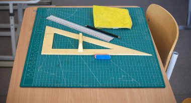 Sınıf masasında cetvel ve kare gibi matematik araçları sergilenirken, bir silgi ve bir kesme paspas üzerindeki temizlik beziyle birlikte.