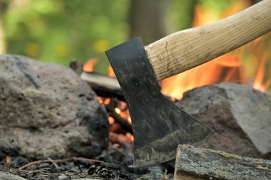 Ahşap saplı bir baltanın yakın görüntüsü, arka planda bir kamp ateşinin sıcacık parıltısı, hayatta kalma ya da tarihi eğlence, boş zaman aktiviteleri ve ateşin etrafında rahatlama.