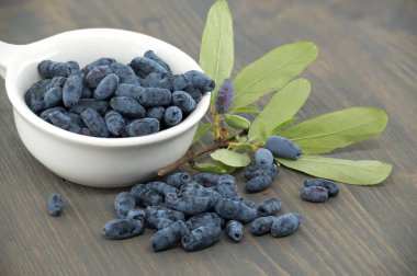 Borealis Honeyberry böğürtlenleriyle dolu beyaz porselen kase ahşap bir masanın üzerinde, taze yeşil yapraklar ve böğürtlenlerle dolu bir masanın yanına dökülen böğürtlenler.