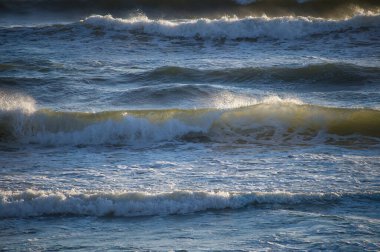 Dynamic ocean waves crash onto a sandy shore at sunset, capturing the beauty and power of nature in a dramatic coastal scene. clipart