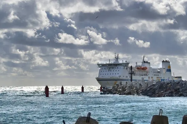 stock image A large ferry navigates through a harbor, highlighted by dramatic, cloudy skies and a breakwater, illustrating marine transportation and infrastructure on a stormy day.