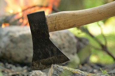 Ahşap bir kütüğe gömülü baltanın yakın görüntüsü, arka planda kamp ateşi, hayatta kalma veya tarihi eğlence, boş zaman aktiviteleri ve ateşin etrafında rahatlama.