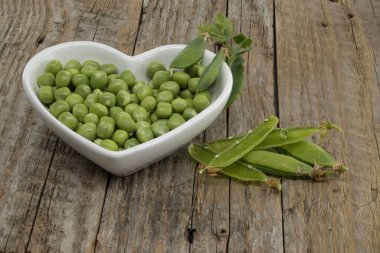 Kırsal ahşap bir masada, kalp şeklinde bir kasede taze bezelyeler kasenin yanında boş bezelye kabukları..
