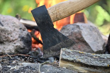 Ahşap bir kütüğe gömülü baltanın yakın görüntüsü, arka planda kamp ateşi, hayatta kalma veya tarihi eğlence, boş zaman aktiviteleri ve ateşin etrafında rahatlama.