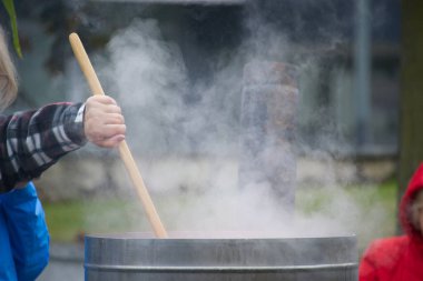 Person stirring large steaming pot outdoors, suggesting communal cooking or food preparation in an industrial-sized metal container clipart