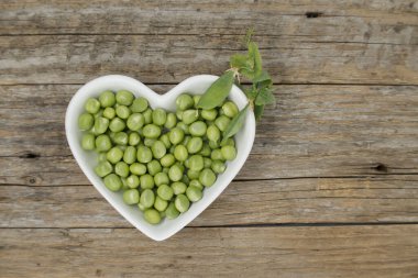 Kırsal ahşap bir masada, kalp şeklinde bir kasede taze bezelyeler, kabın yanında bezelye kabukları..