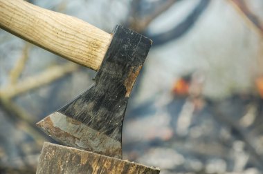 Ağacın kütüğüne saplanmış balta, arka planda yanan kamp ateşi, odun kesmek, odun kırmak veya çevresel kaynak yönetimi gibi açık hava aktiviteleri.