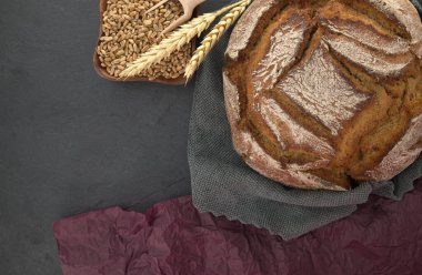 Ekmek somunu ve buğday tanecikleri gri bir mutfak havlusunun üzerine siyah bir karatahtaya yerleştirilmiş.