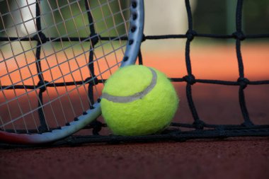 Tenis topu ve raket, gün batımında kil kortun ağında dinleniyor. Rekabetin heyecanıyla eğlenceyi harmanlayan büyüleyici bir atmosfer yaratıyor.