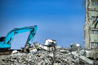 Açık havada, büyük bir binanın yıkılması sırasında mavi gökyüzü arka planına sahip bir moloz yığınının üzerinde bir deniz mavisi kazıcı duruyor..