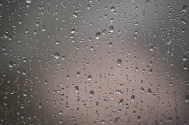Bulanık bulutlu bir günde, farklı boyutlarda yağmur damlalarıyla kaplı pencerenin yakın plan görüntüsü. Kompozisyon, su damlacıklarının dokularını ve soyut desenleri vurguluyor, sakin iç gözlemleri çağrıştırıyor